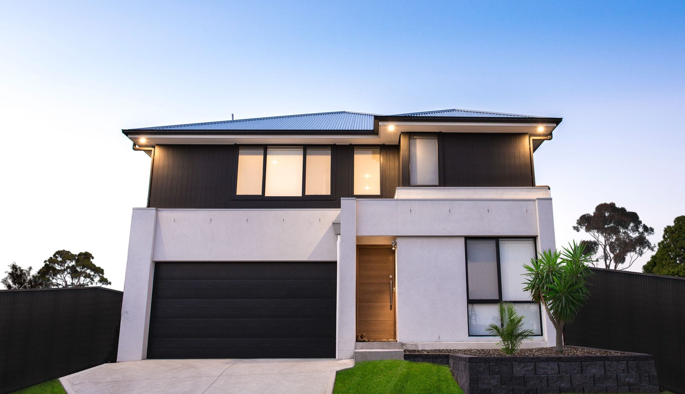 Two-storey rendered home at dusk with charcoal cladding, timber front door and lit garage forecourt